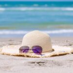 Ein Strohhut und eine Sonnenbrille, die auf dem Strand liegen. Im Hintergrund sieht man blaues Wasser.