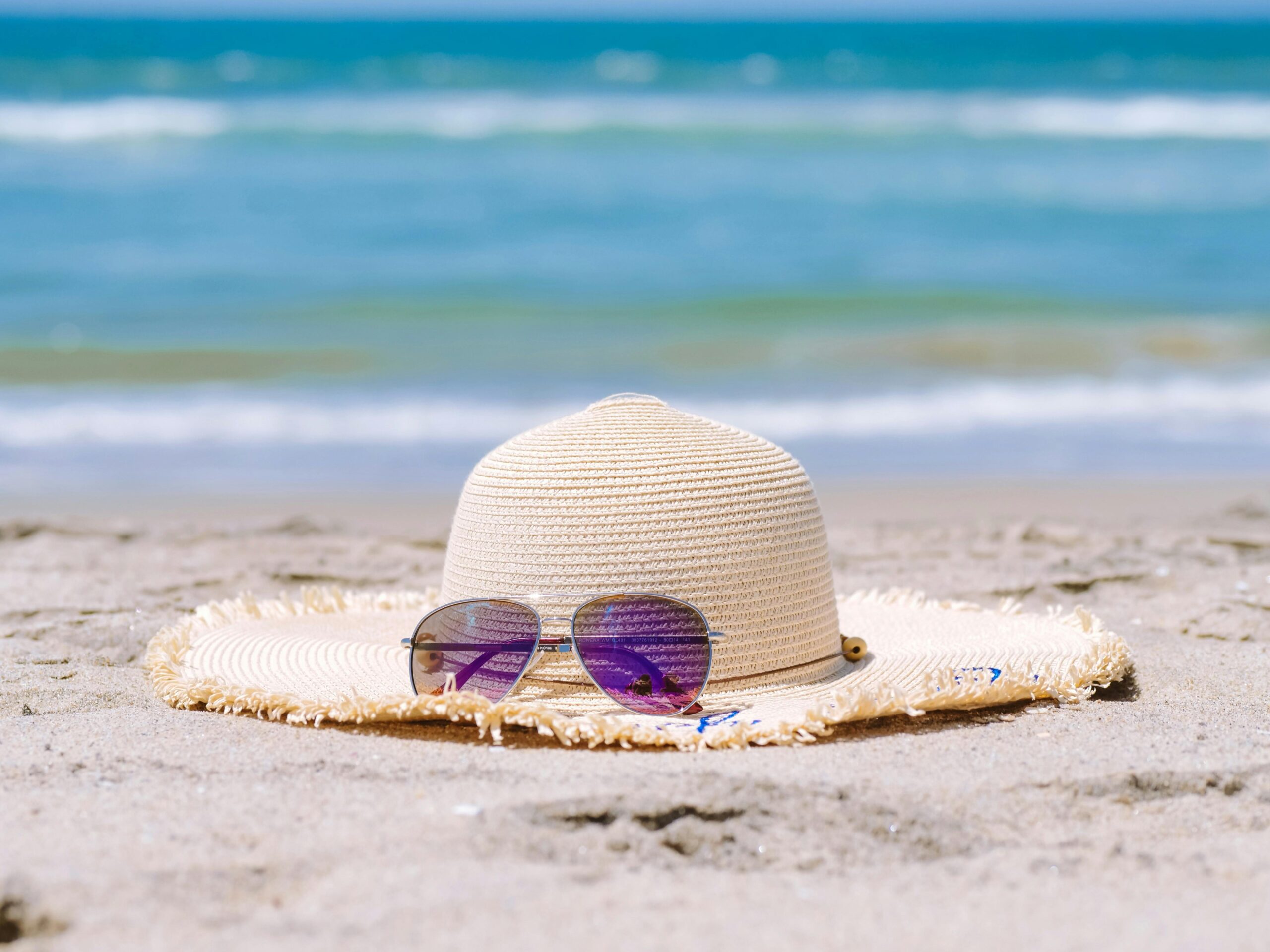 Ein Strohhut und eine Sonnenbrille, die auf dem Strand liegen. Im Hintergrund sieht man blaues Wasser.